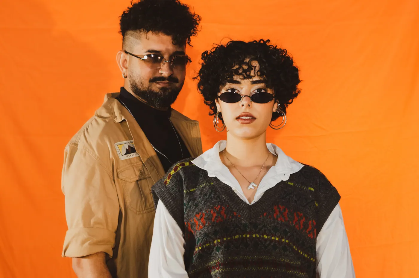Elias e Flavy lado a lado, de óculos escuros, sobre fundo laranja